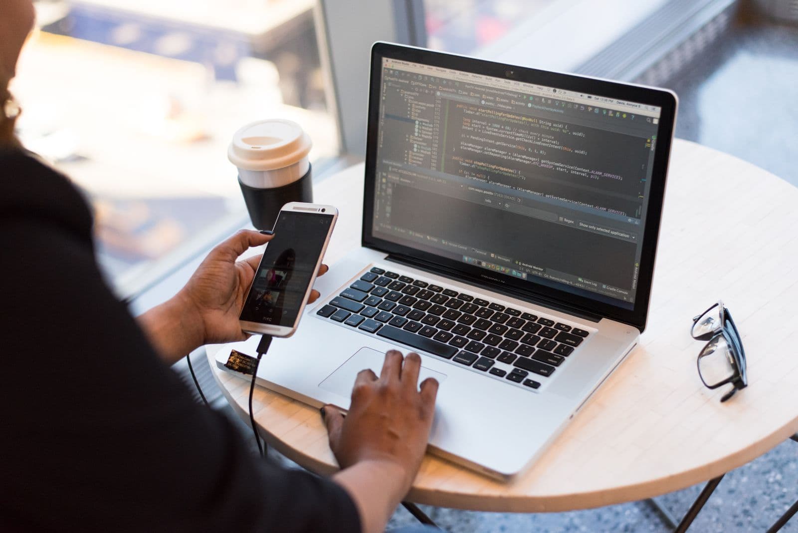 A person working on a laptop showing code at a cafe table, holding a phone beside a coffee cup and glasses