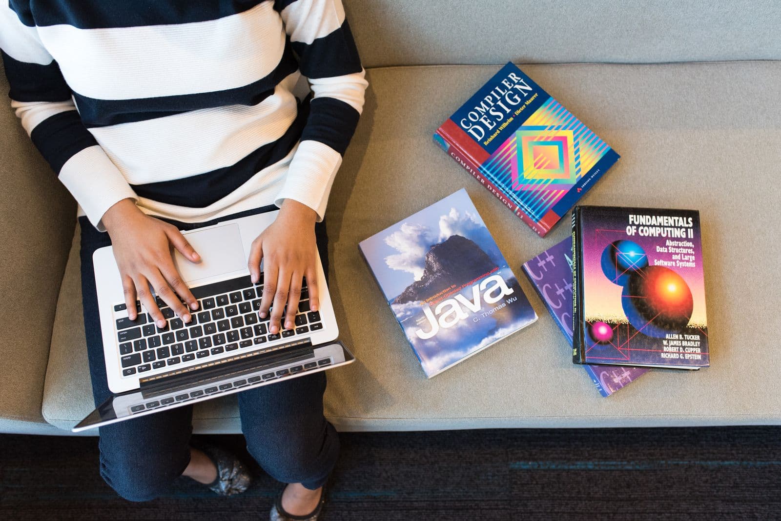 Overhead view of a person typing on a laptop on a grey sofa beside three computing textbooks