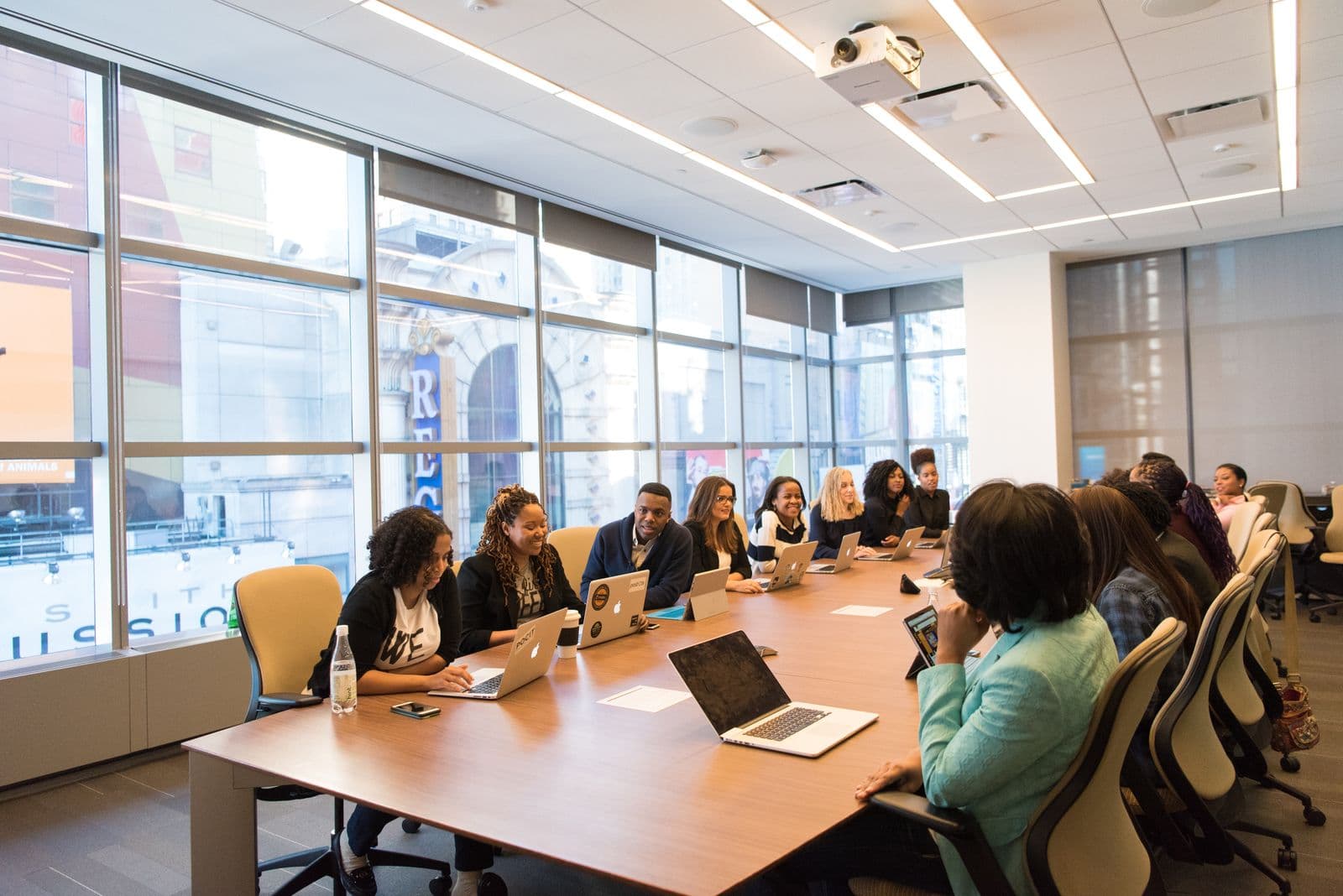 A long boardroom table lined with people at open laptops beside a wall of windows overlooking a city street