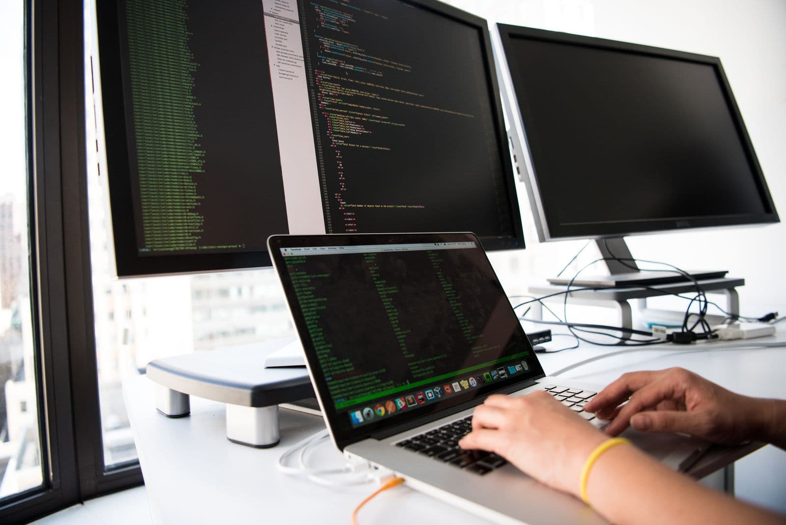 A person typing on a laptop in front of two large monitors displaying dense lines of code beside a bright window