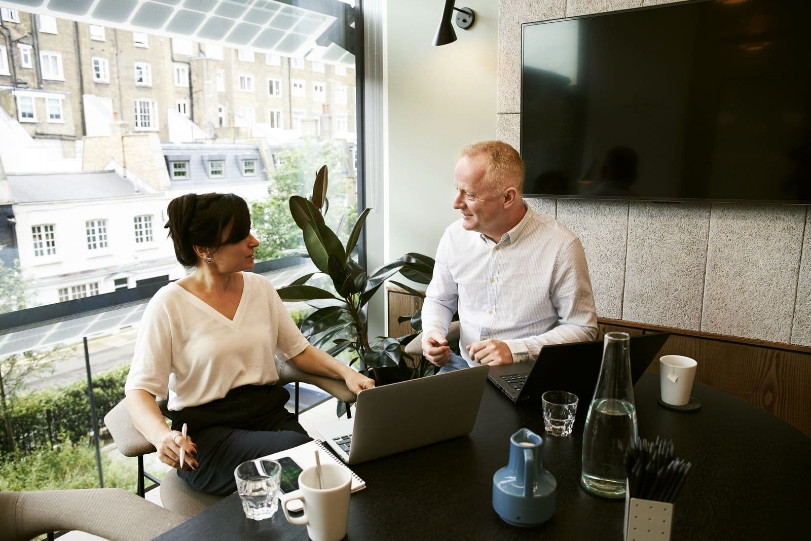 Two colleagues talking across a dark table with laptops, a water bottle and glasses, beside a bright window