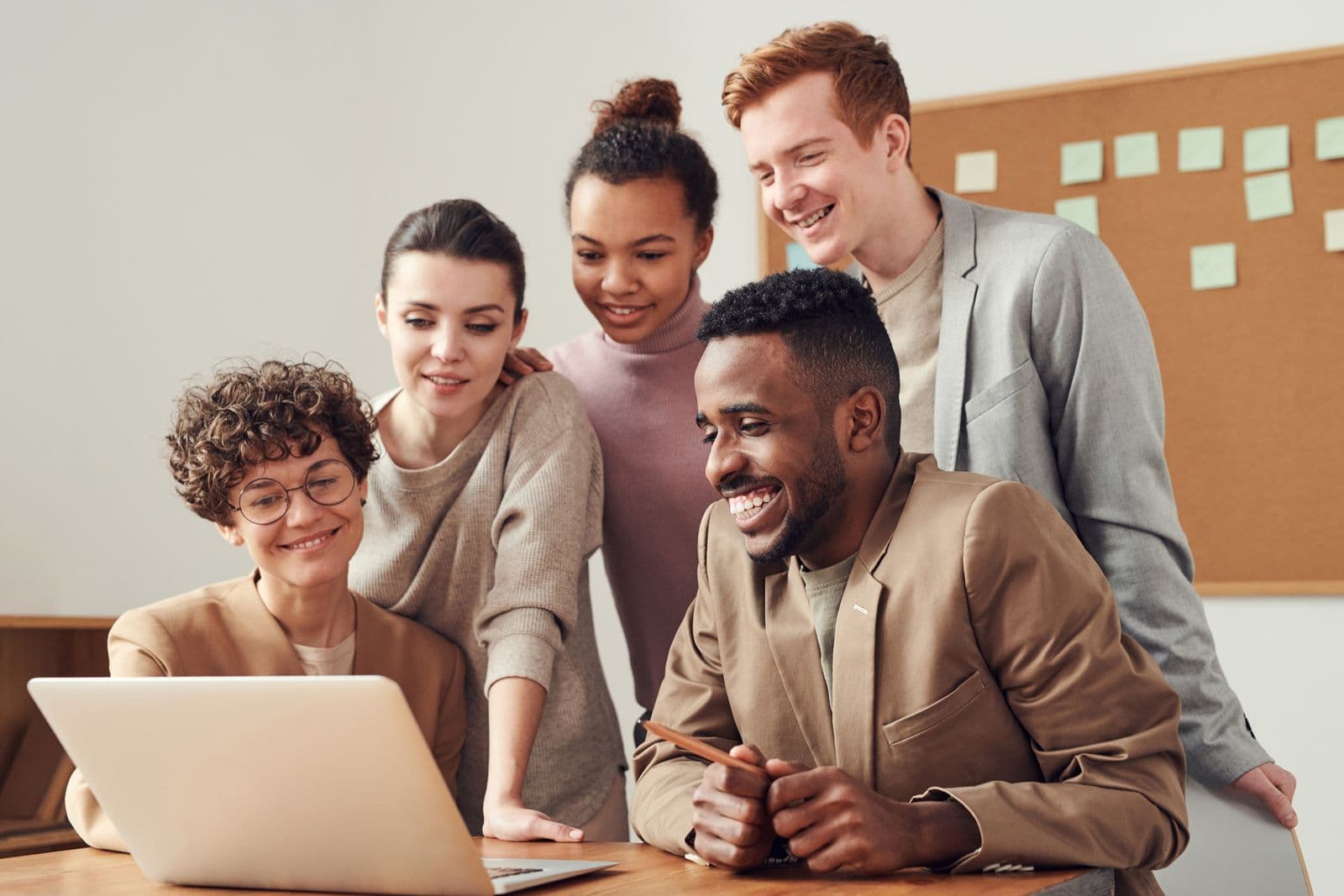 Four colleagues gathered around a seated man at an open laptop, all smiling at the screen in a bright office