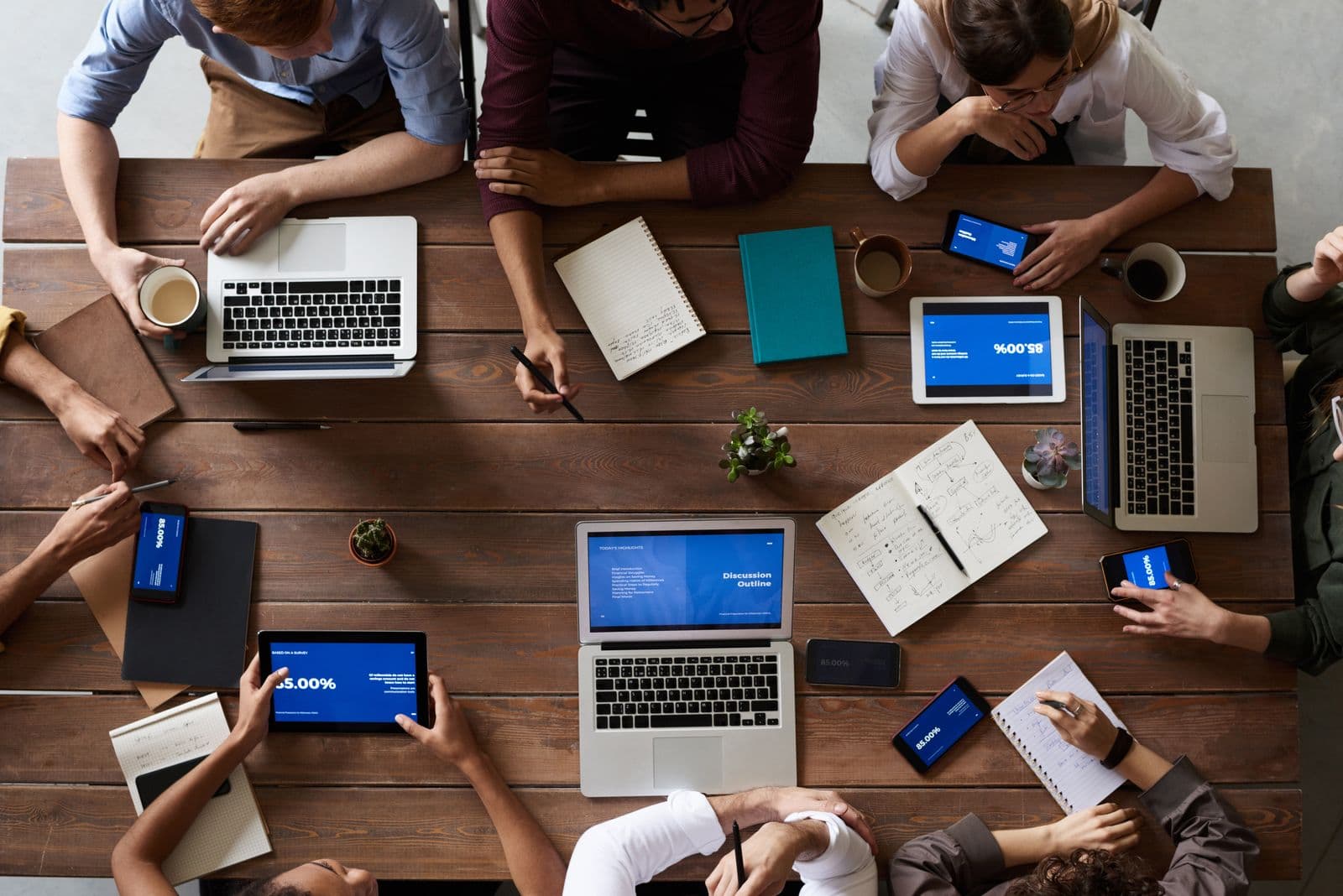 Overhead view of a wooden table ringed by colleagues with laptops, tablets, notebooks and coffee cups