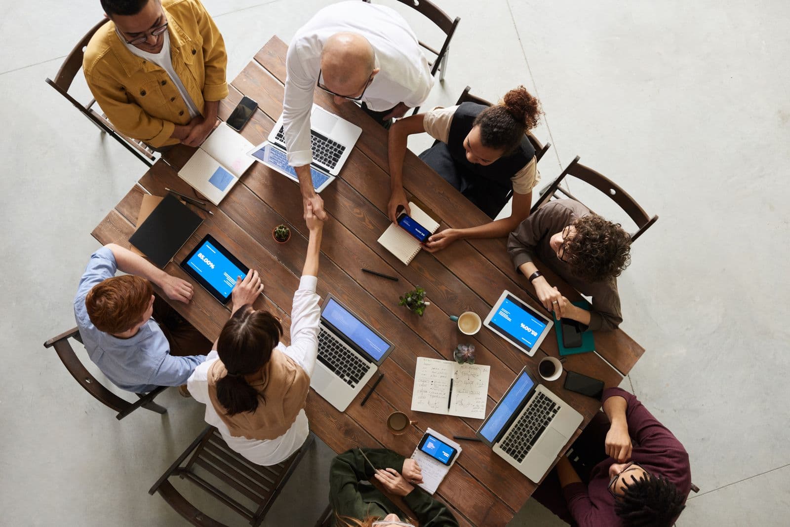 Overhead view of eight colleagues seated around a large wooden table with laptops, tablets and notebooks
