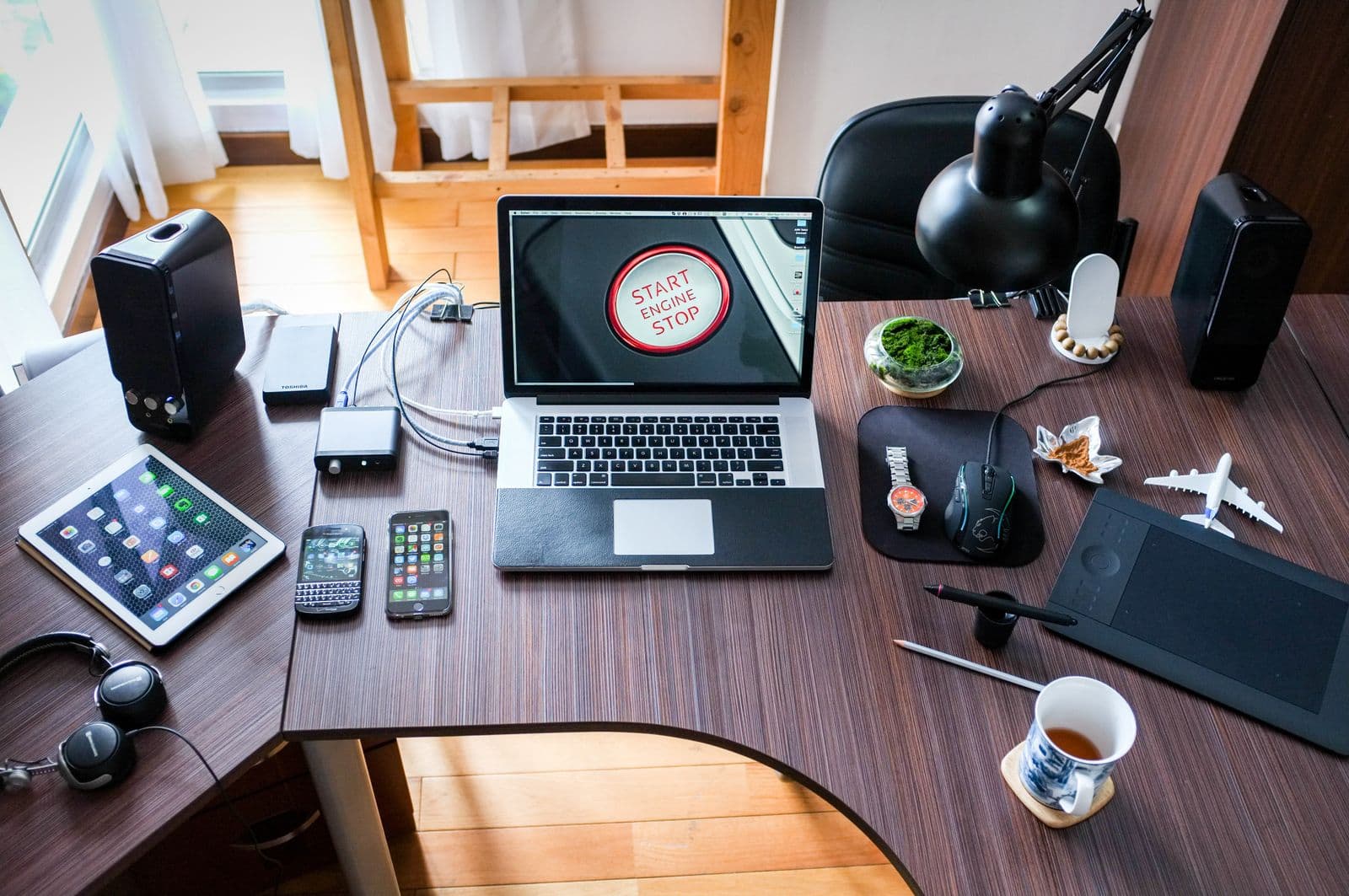 A wooden desk with a laptop, tablet, two phones, headphones, speakers and a drawing tablet arranged neatly