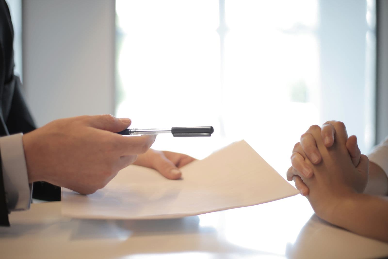 One person offering a pen across a white table to another whose hands are clasped over a contract