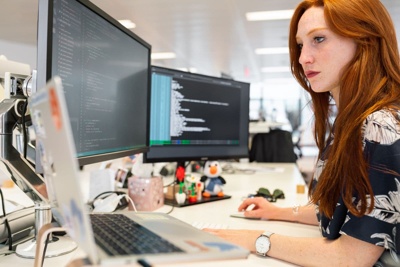 A developer working at a desk with two large monitors displaying code in an open-plan office