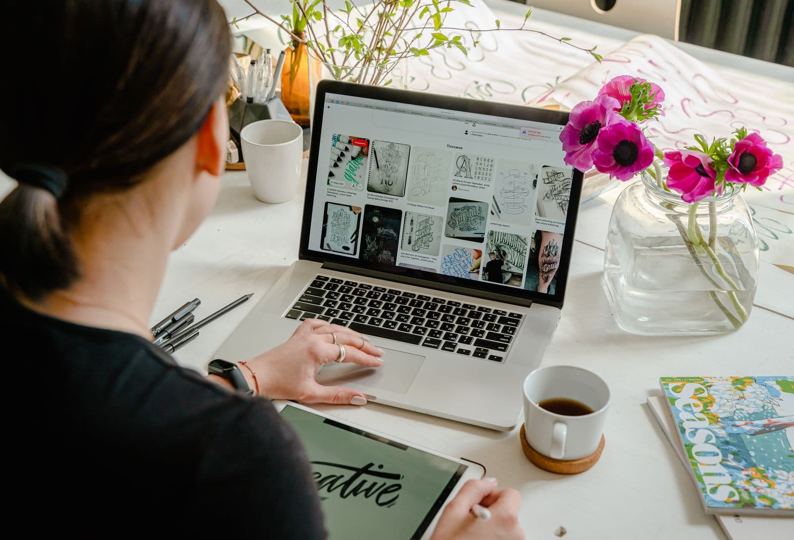 A woman at a laptop reviewing a grid of sketches on screen, with a graphics tablet, coffee and anemones on the table