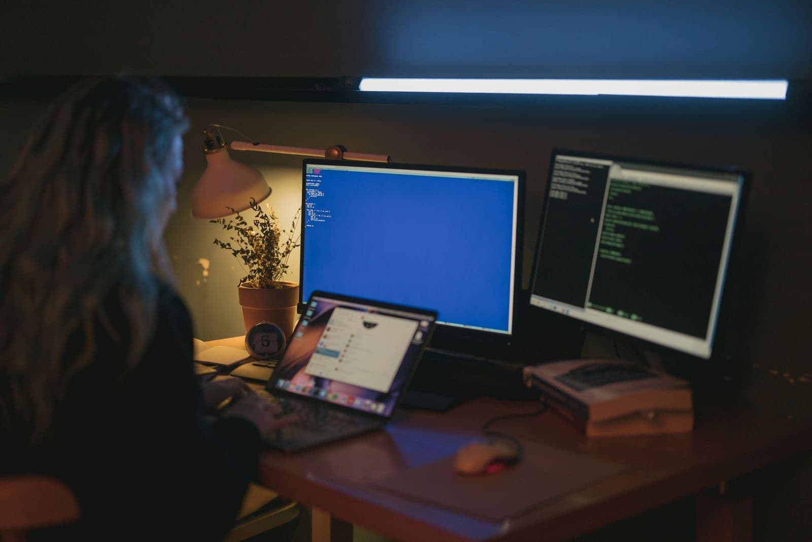 A person working in a dim room at a desk with a laptop and two monitors, one showing code and one glowing blue