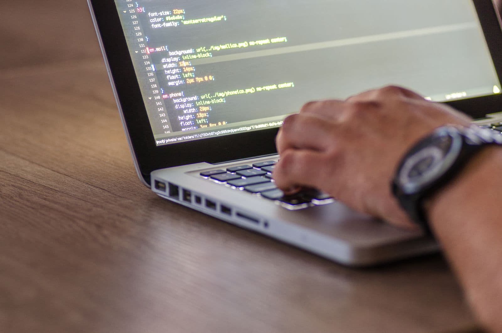 A hand resting on a laptop keyboard on a wooden desk, the screen showing coloured lines of stylesheet code