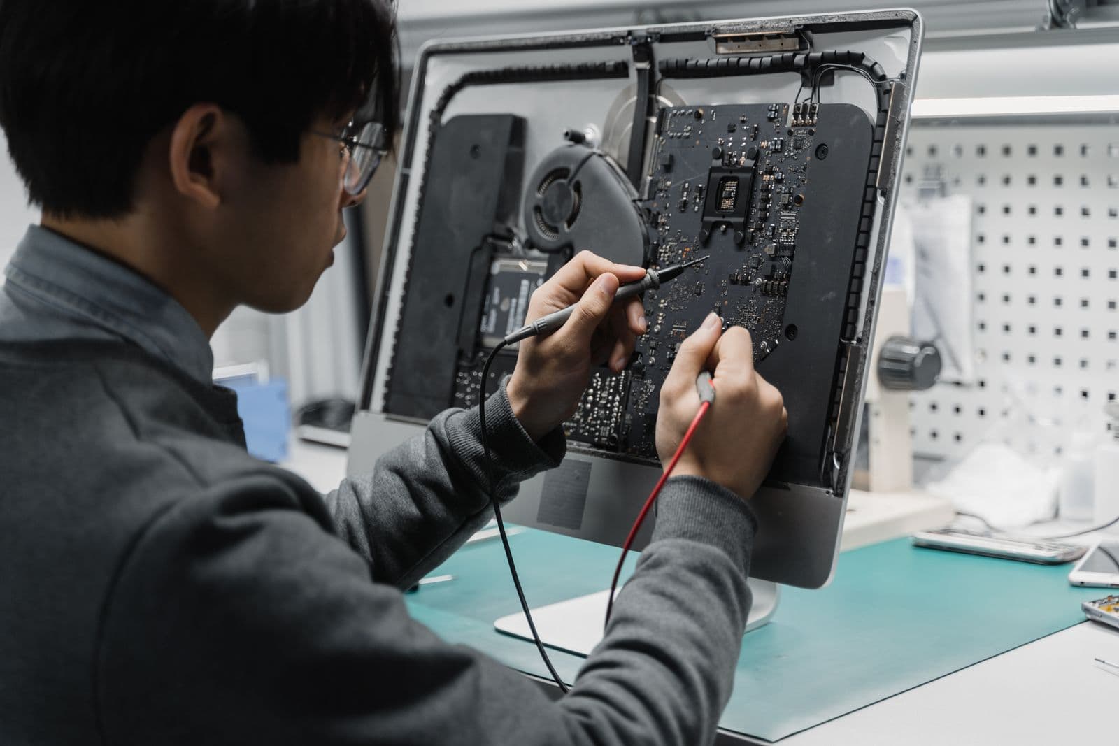 Technician in glasses using multimeter probes to diagnose the circuit board of an opened all-in-one desktop computer at a repair bench