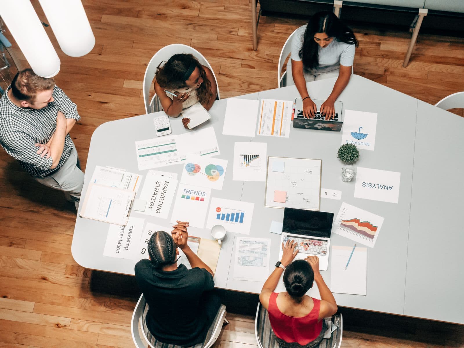 Overhead view of five colleagues at a white table covered with printed charts labelled marketing strategy and analysis