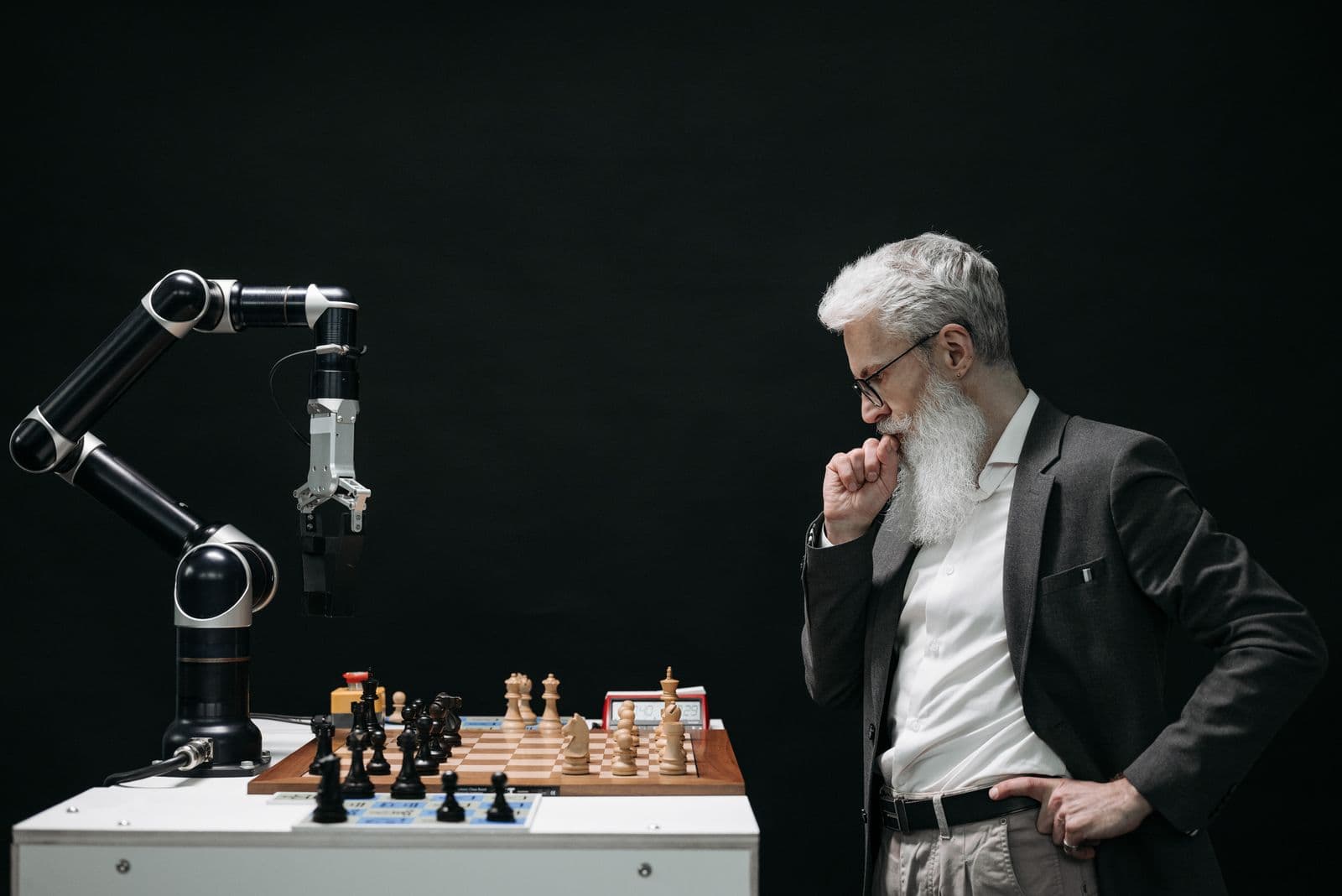 A bearded man in a blazer studying a chessboard across from a black robotic arm poised over the pieces